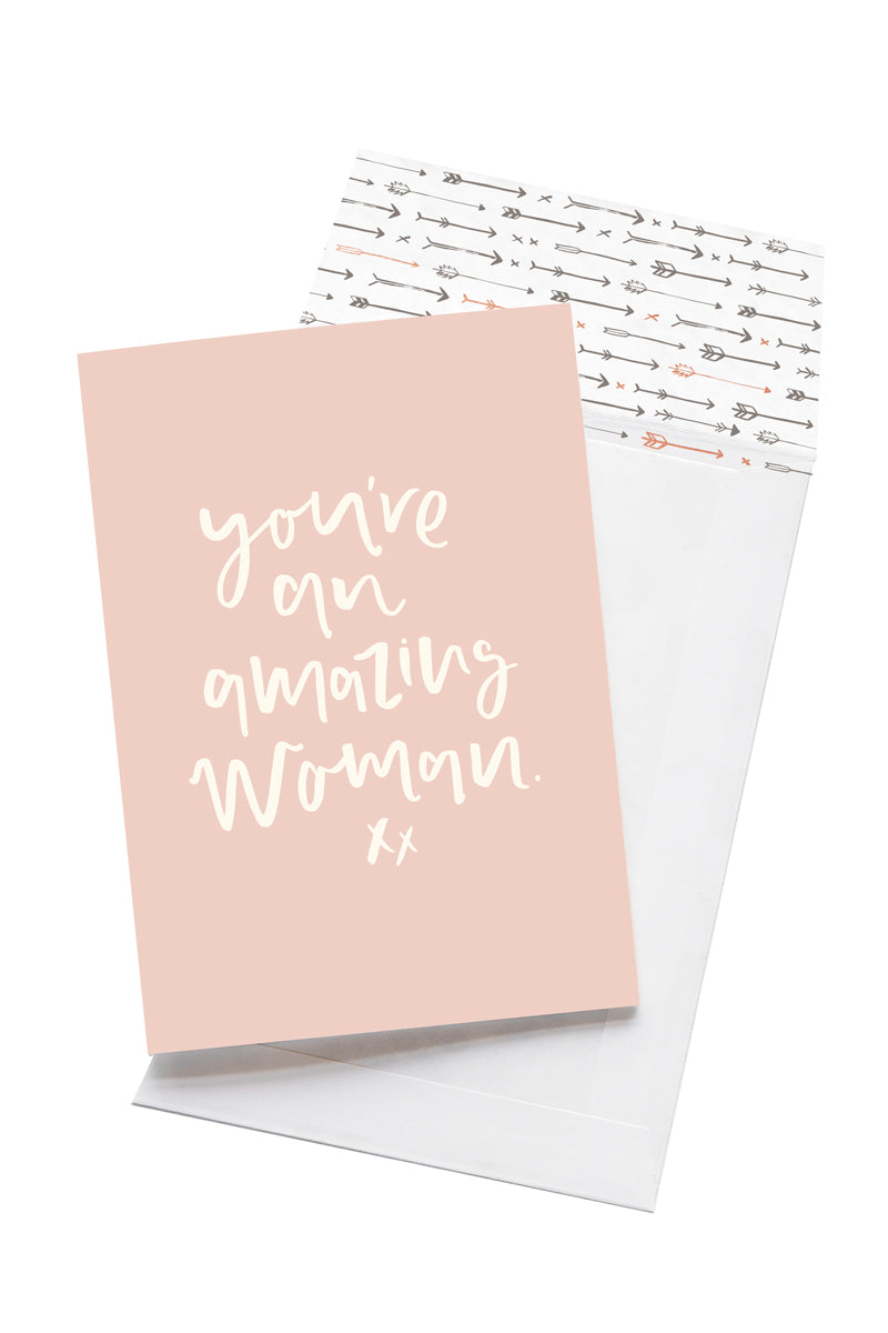 A pale pink card with the message 'you're an amazing woman. xx' is sitting on a white background. The card uses white script font. There is a white envelope behind the card which has an arrow pattern design inside.