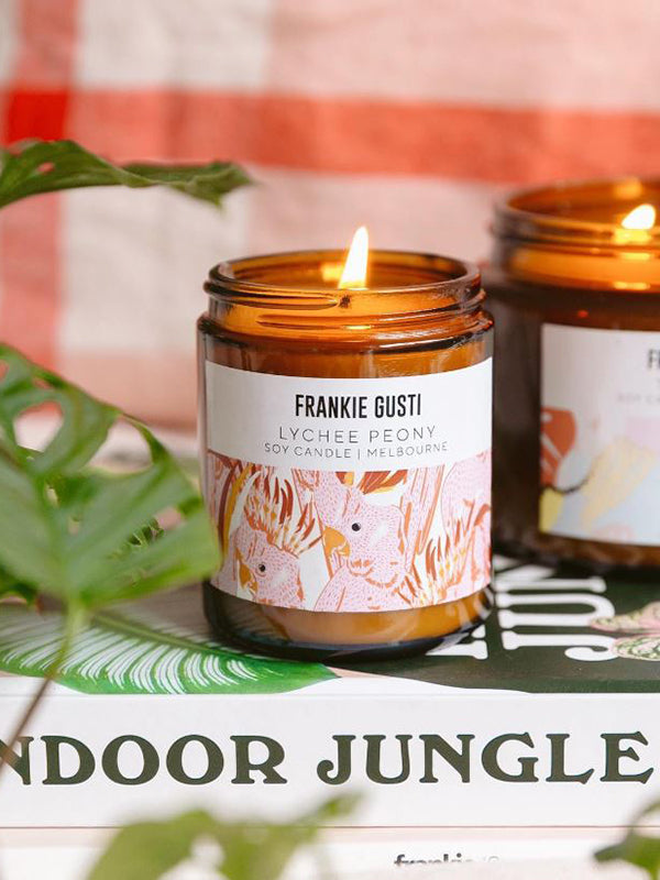 A Frankie Gusti Lychee Peony candle is styles against a leafy green background. The candle is lit, and another candle is blurred in the background. The text Indoor Jungle is written below the candle in green.