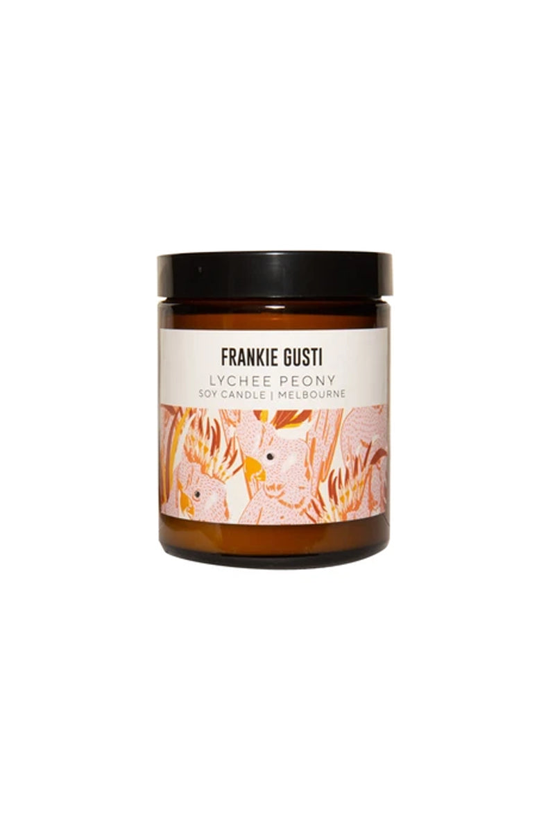 A candle jar sits against a white background. The jar is amber and has a black lid, it has a label with artwork featuring peach and yellow cockatoos. It has a cream box up the top with the words FRANKIE GUSTI LYCHEE PEONY SOY CANDLE | MELBOURE written in modern uppercase text.