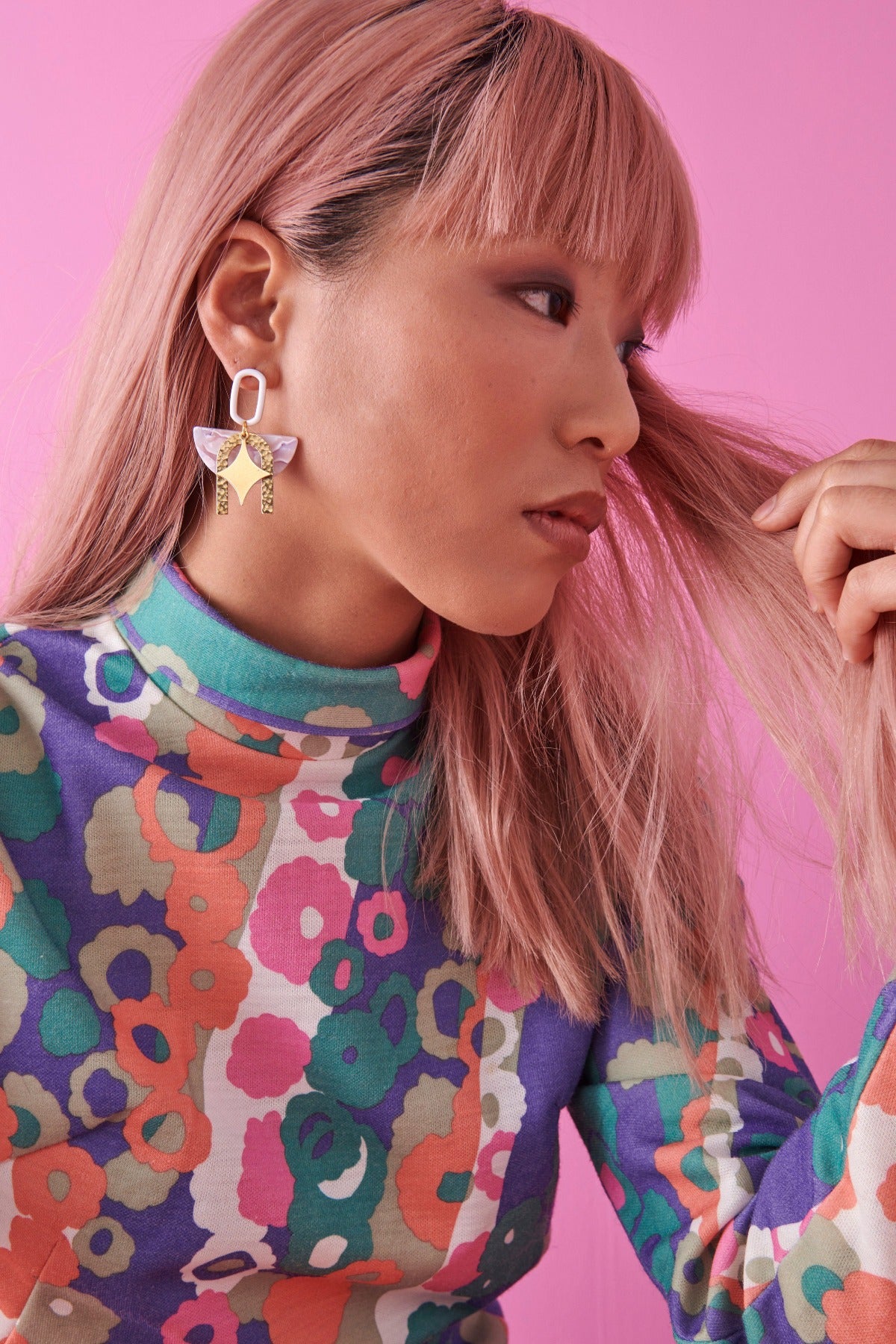 A pink haired lady shows a side view of the white Beatnik earring. She wears a graphic floral dress and is seated against a pink background. 