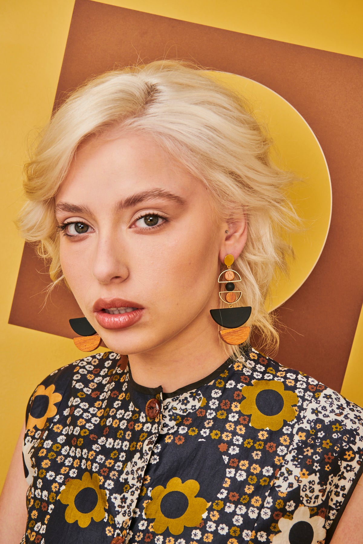 A lady with short blonde hair sits against a yellow and brown background. She models the Marcel earrings in black and wears a black, tan and mustard retro floral print top.