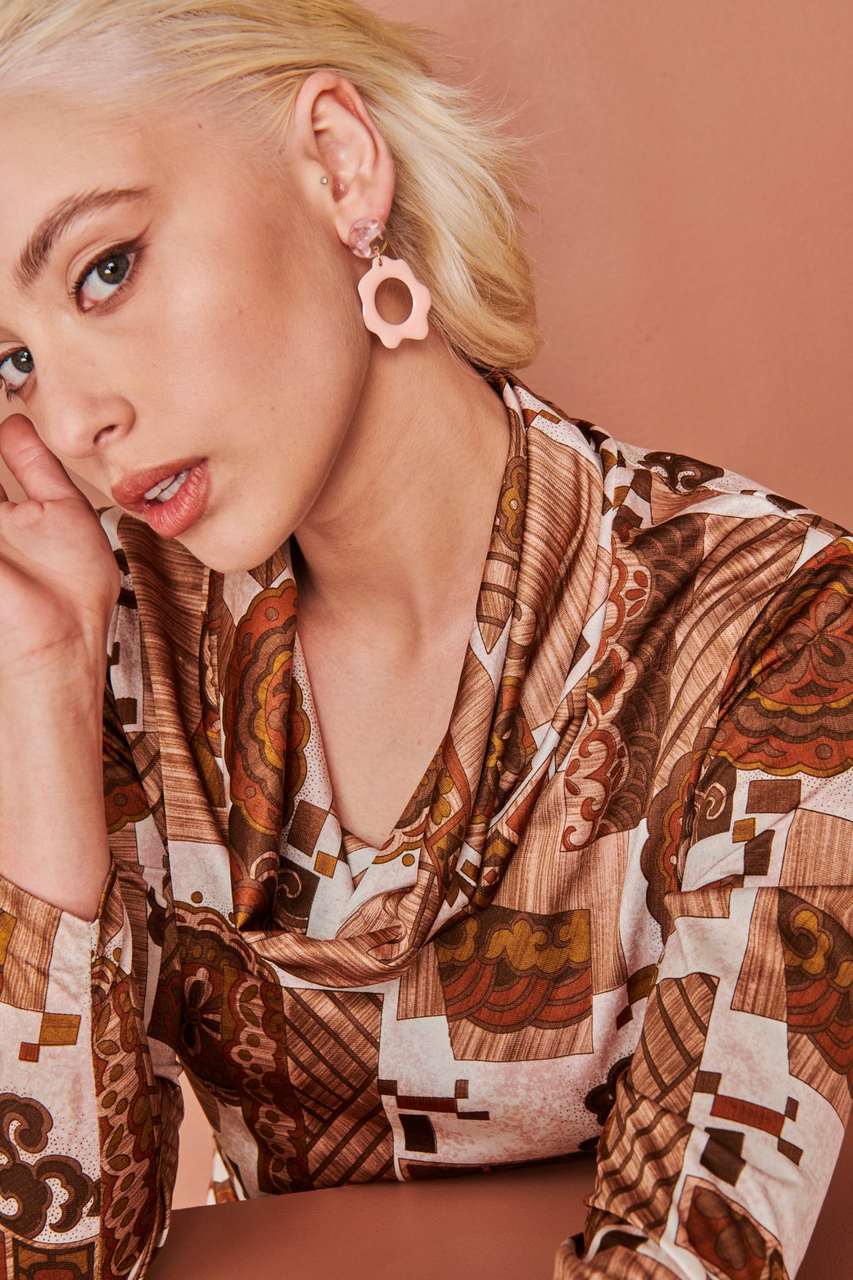 A lady with blonde hair sits against a peach background. She models a side-view of the Floret earrings in marshmellow and wears a brown retro print top with a cowl neckline.