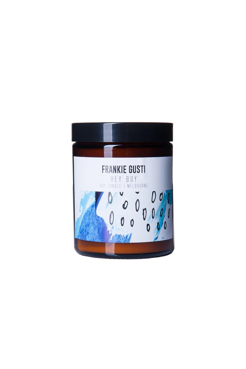 A candle jar sits against a white background. The jar is amber and has a black lid, it has a label featuring abstract art in navy, white and black. It has a white box up the top with the words FRANKIE GUSTI HEY BOY SOY CANDLE | MELBOURE written in modern uppercase text.