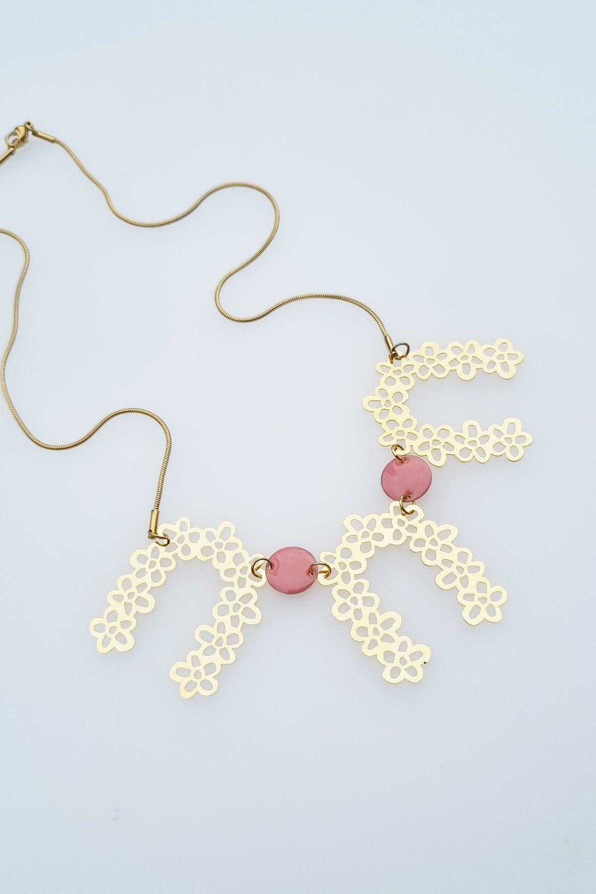 A necklace sits against a white background. It features a gold chair with three floral brass arch pieces, and two pink enamel connector dots separating each arch.