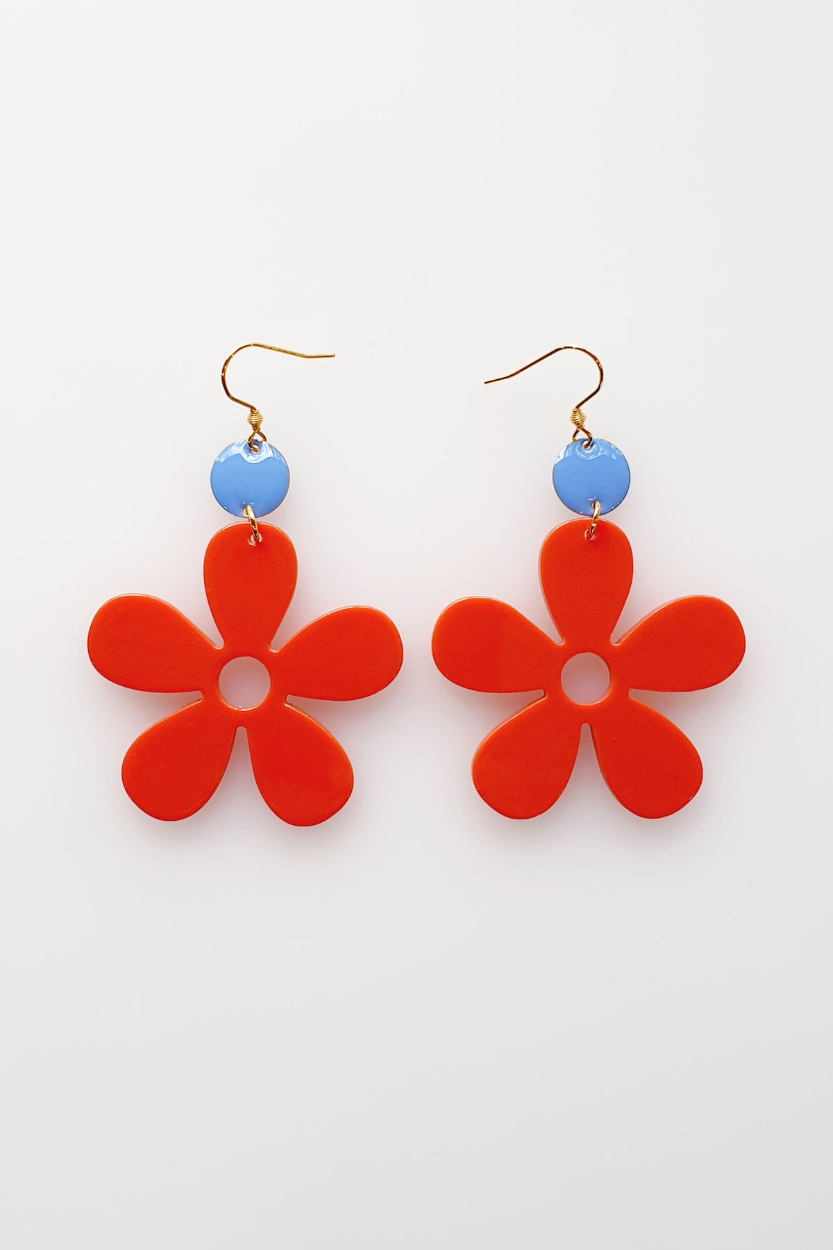 A pair of red acrylic daisy dangle earrings with a blue enamel dot attached to a hook. Pictured against a white background.