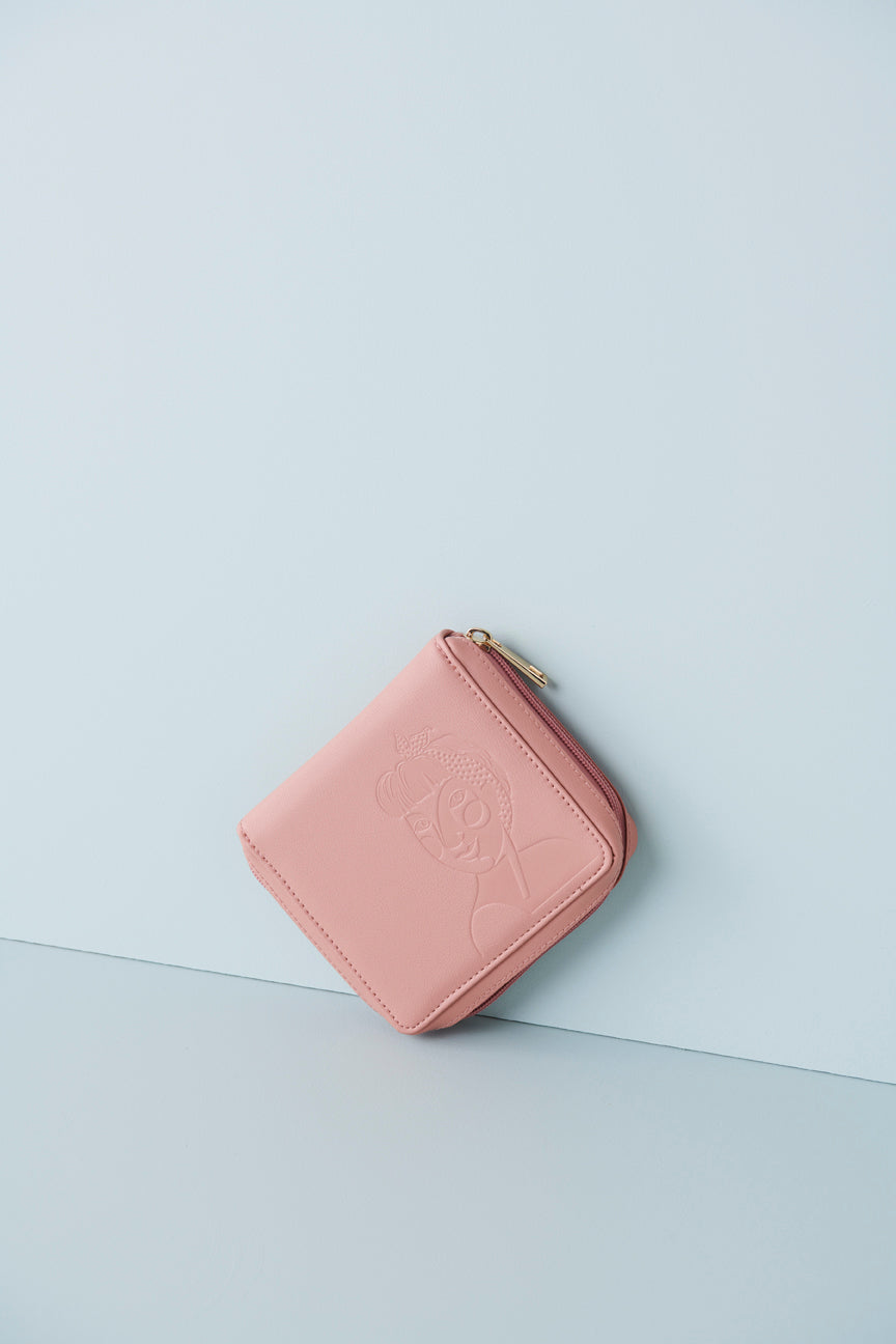 A pink jewellery wallet sits on an angle against a baby blue background. The wallet features an embossed image of a lady with a fringe and head scarf. It has a pink zip and has a gold zip pull.
