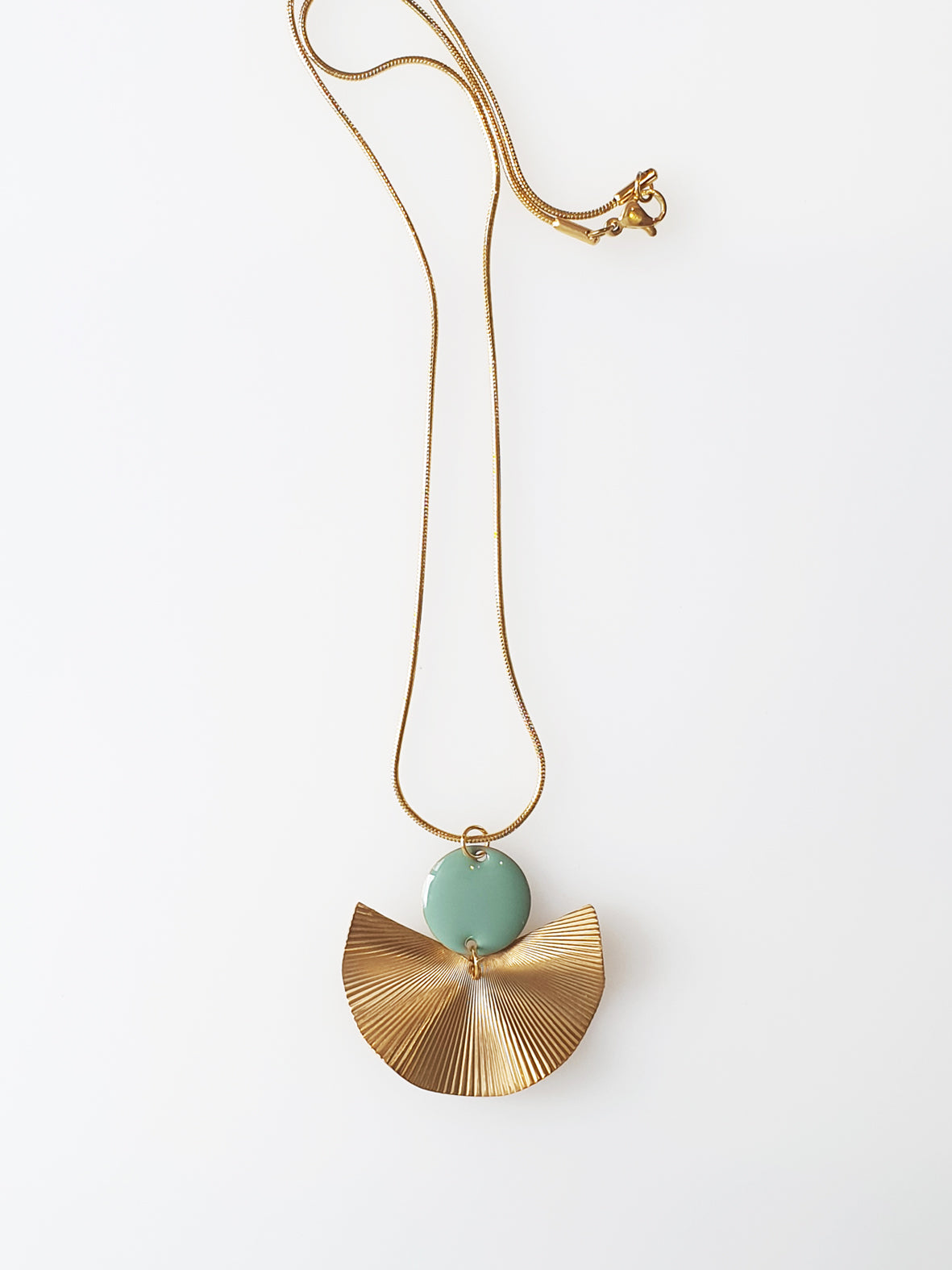 A necklace sits against a white background. It features a gold chain, a duckegg enamel connector dot and a textured brass fan piece.