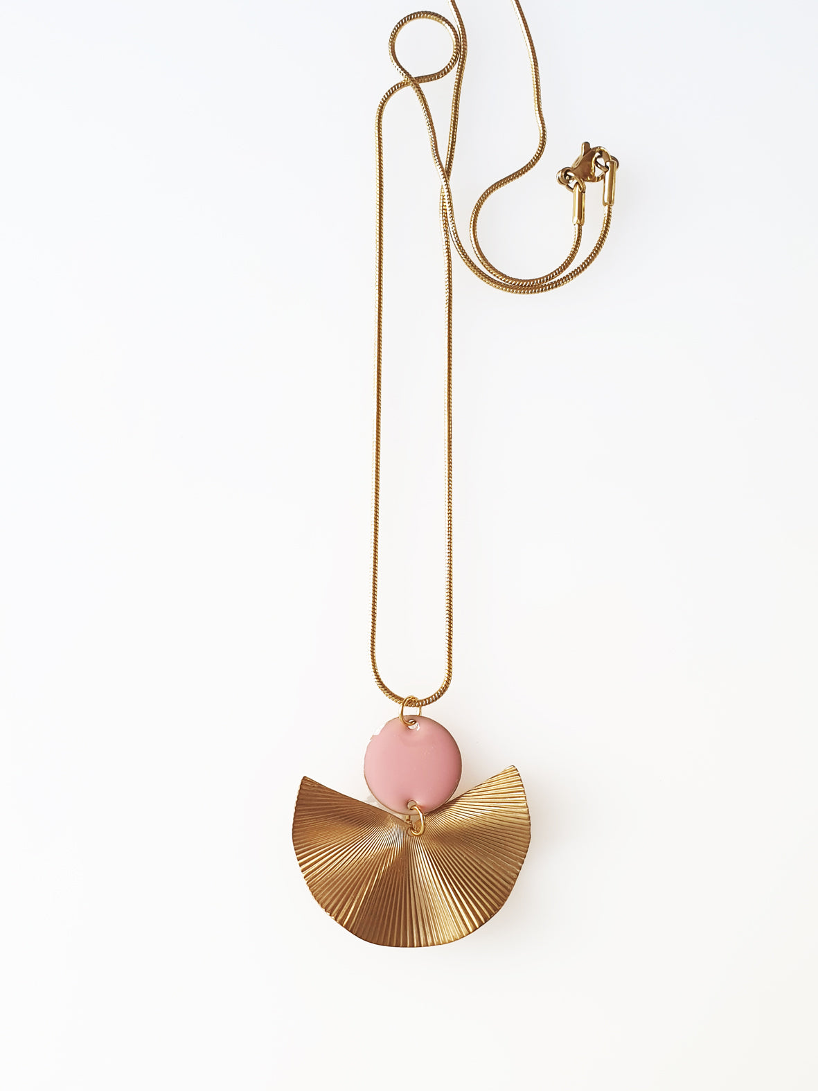 A necklace sits against a white background. It features a gold chain, a pink enamel connector dot and a textured brass fan piece.