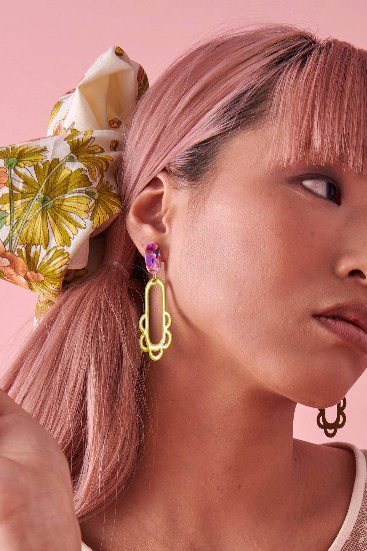 A lady with pink hair models a sideview of the Fiesta earrings in avocado. She sits against a pink background and wears a floral scarf tied through her ponytail.