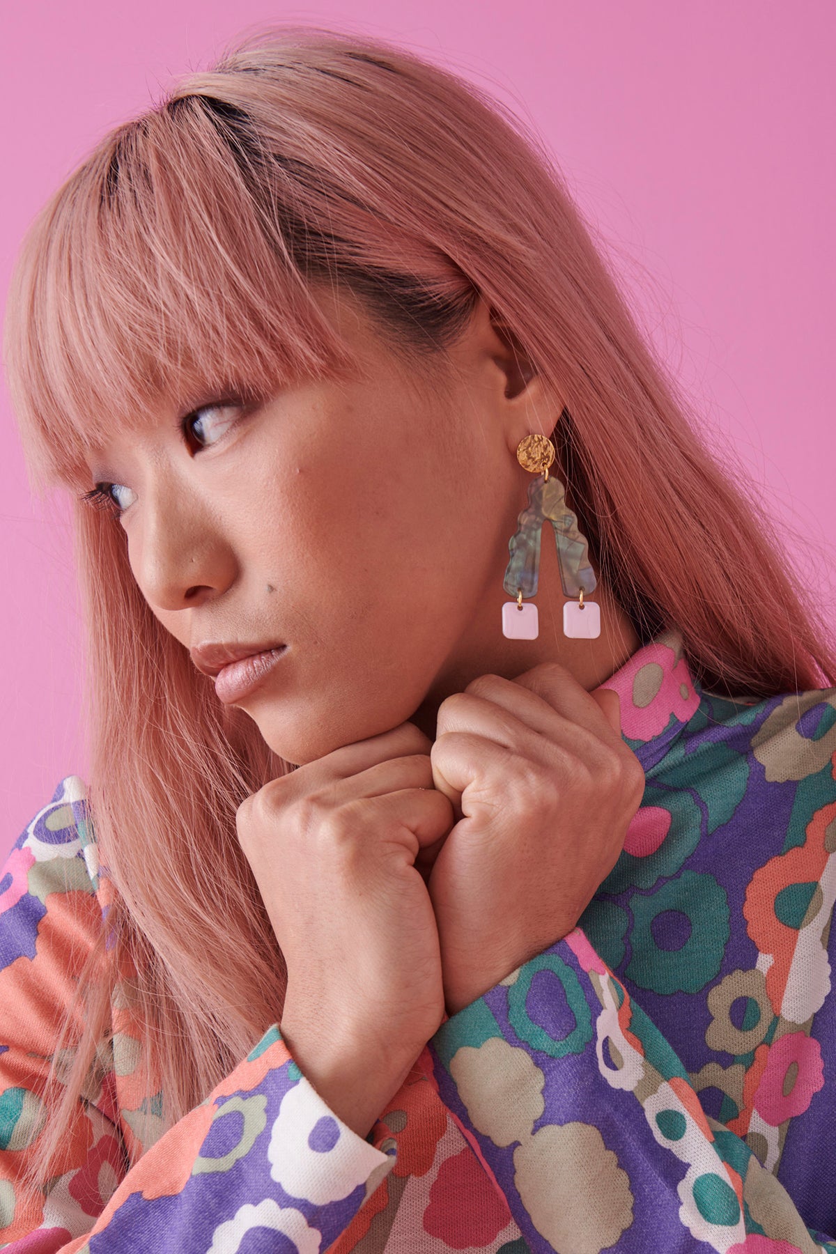A lady with pink hair sits against a bright pink background. She models a sideview of the Groove Thang earrings in green. Her hands are closed in fists and tucked under her chin and she wears a retro multicoloured floral top.