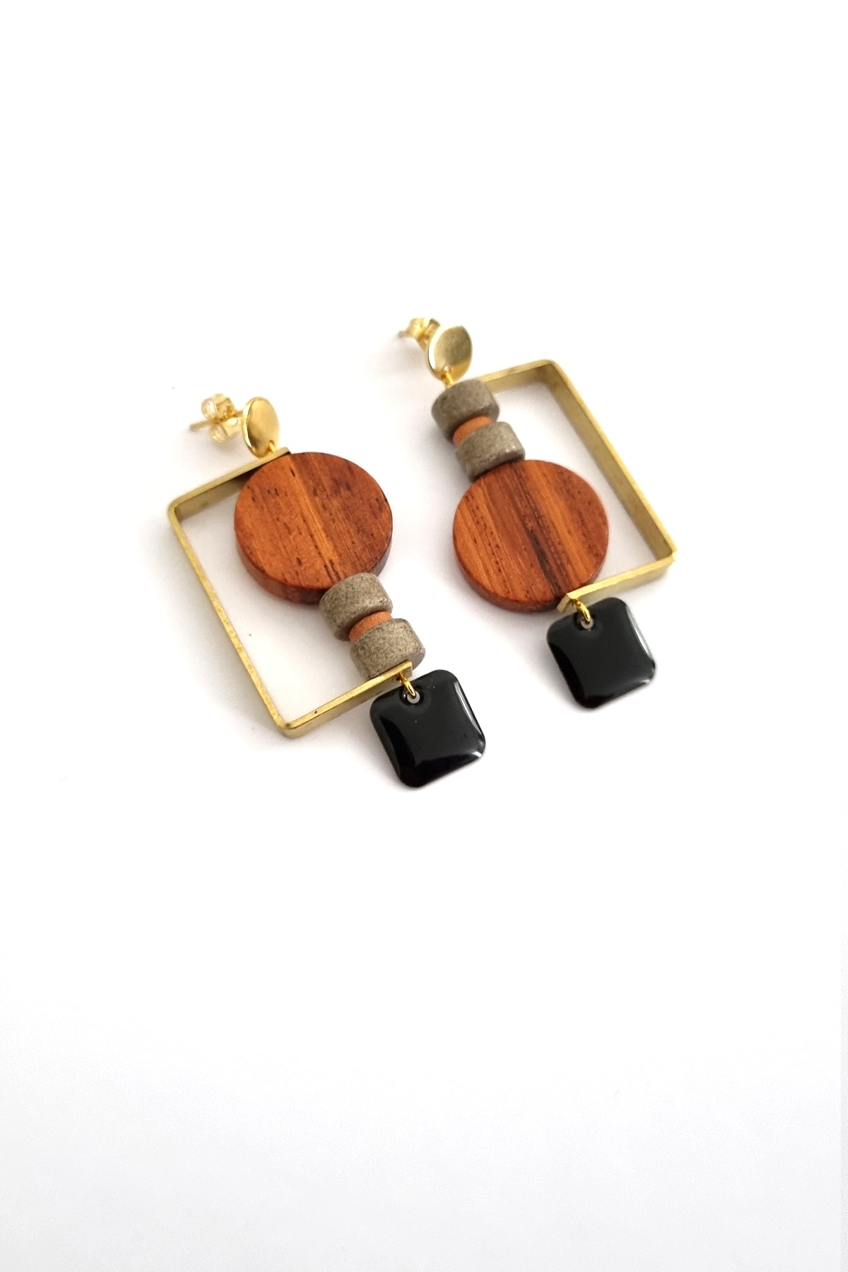 A pair of stud dangle earrings lay against a white background. They feature a half square brass shape that envelopes a circular wooden bead, and two grey stone beads separated by a small wooden bead. The design is flipped in each ear and a black square enamel dangle hangs from the bottom on each earring.