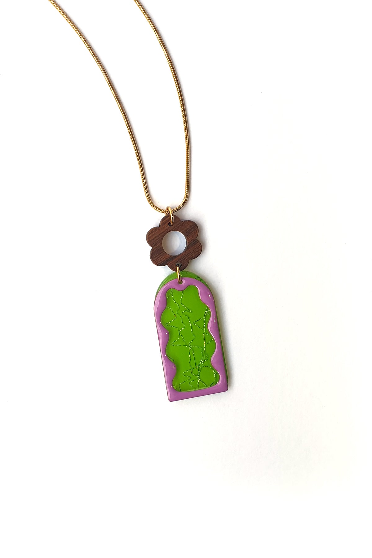 A necklace lays against a white background. It features a gold chain, a wooden flower top, followed by a green acrylic arch with silver thread detail overlaid by a purple acrylic wavy arch frame.