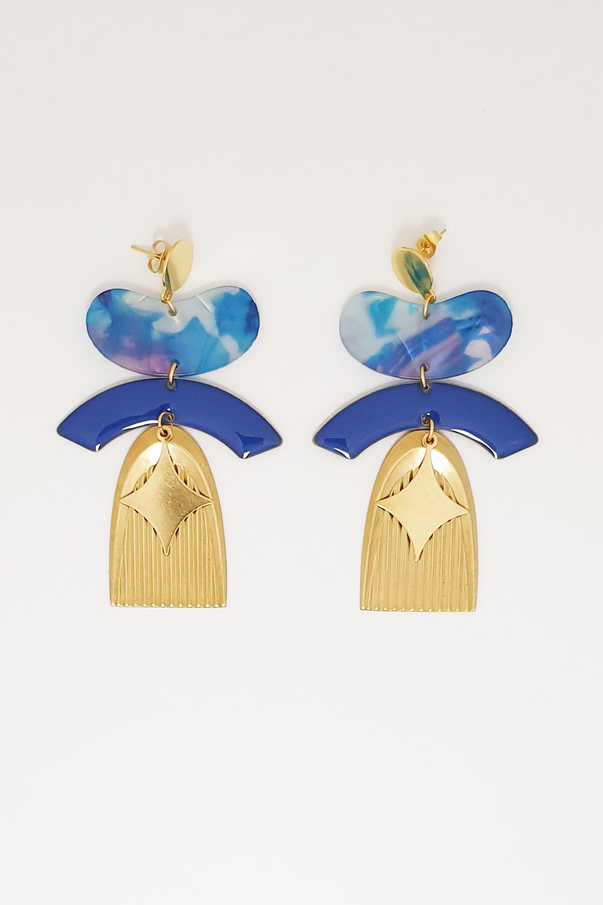 A pair of statement stud dangle earrings sit against a white background. They feature a sapphire blue jellybean shaped acrylic piece, a sapphire blue arch shaped enamel connector, a corrugated textured brass arch piece, and a diamond shaped brass piece.