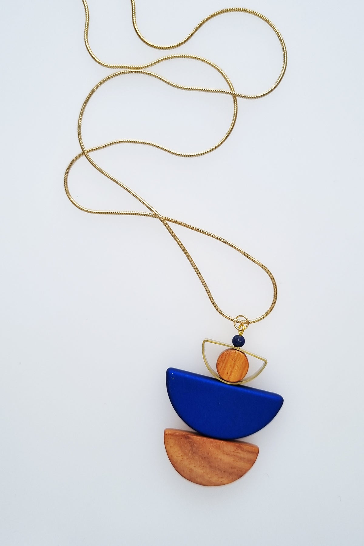 A necklace with a gold chain sits against a white background. It features a small wooden bead, followed by a brass D shape with a wooden circle bead enclosed, followed by a blue D shaped bead, and a wooden D shape bead.