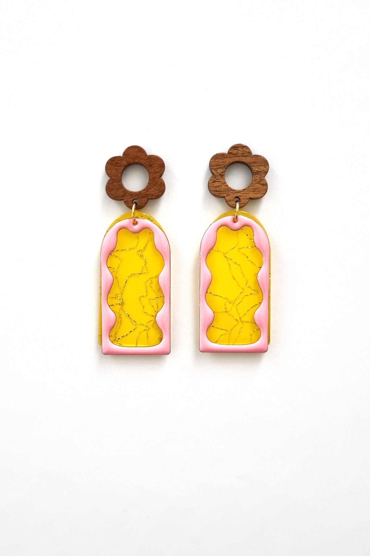 A pair of stud dangle earrings lay against a white background. They feature a wooden flower stud top followed by a yellow acrylic arch with a silver thread detail. On top sits an enamel wavy arch frame in pink.