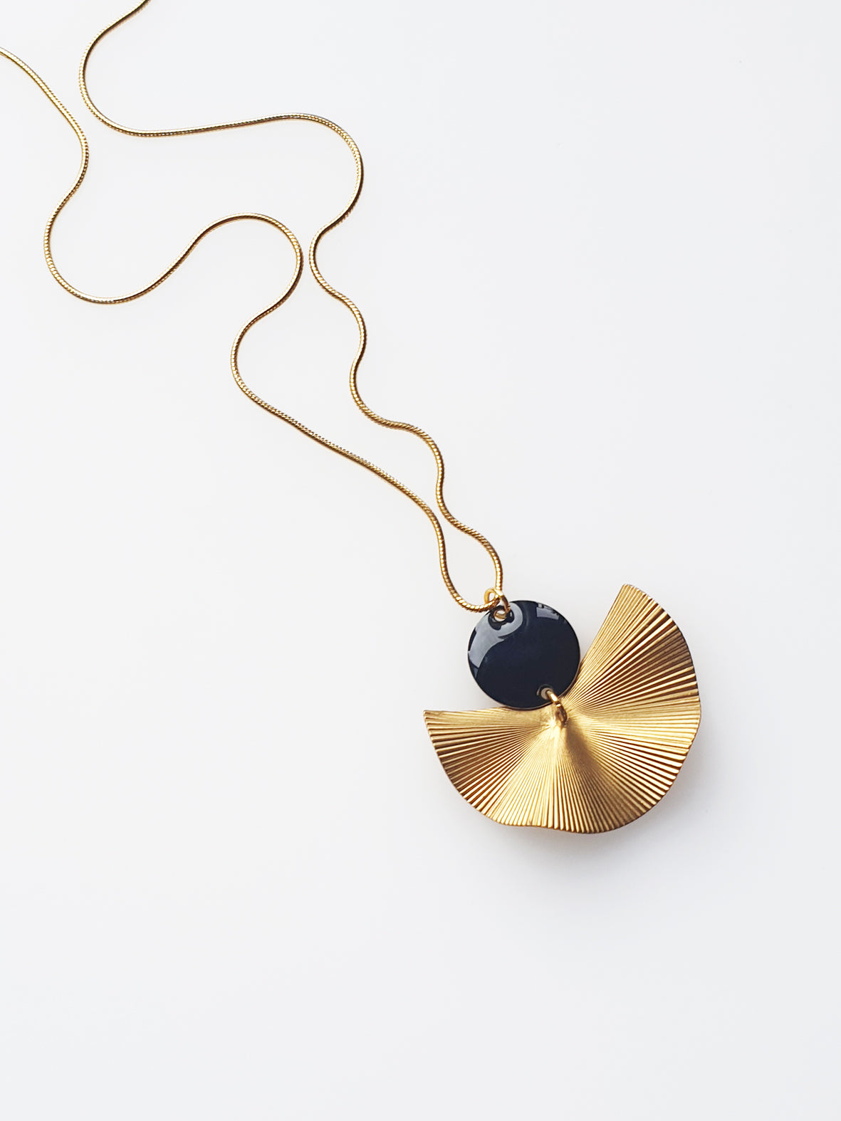 A necklace sits against a white background. It features a gold chain, a black enamel connector dot and a textured brass fan piece.
