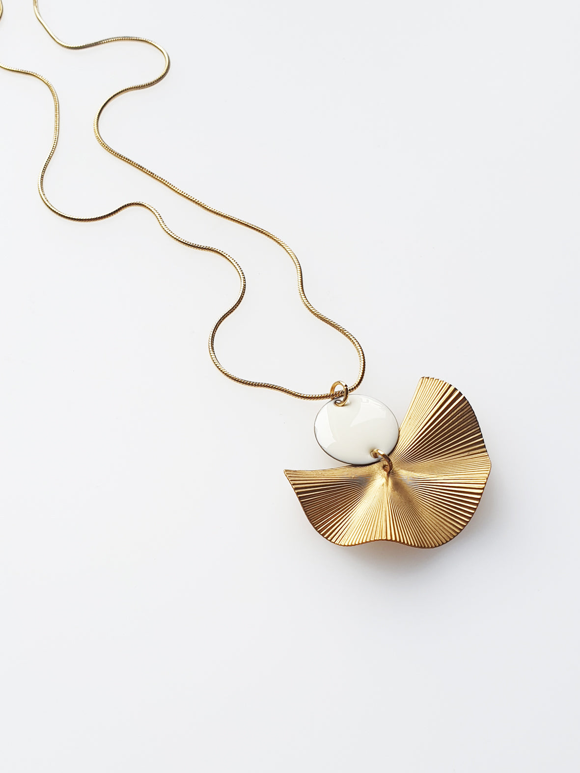 A necklace sits against a white background. It features a gold chain, a white enamel connector dot and a textured brass fan piece.