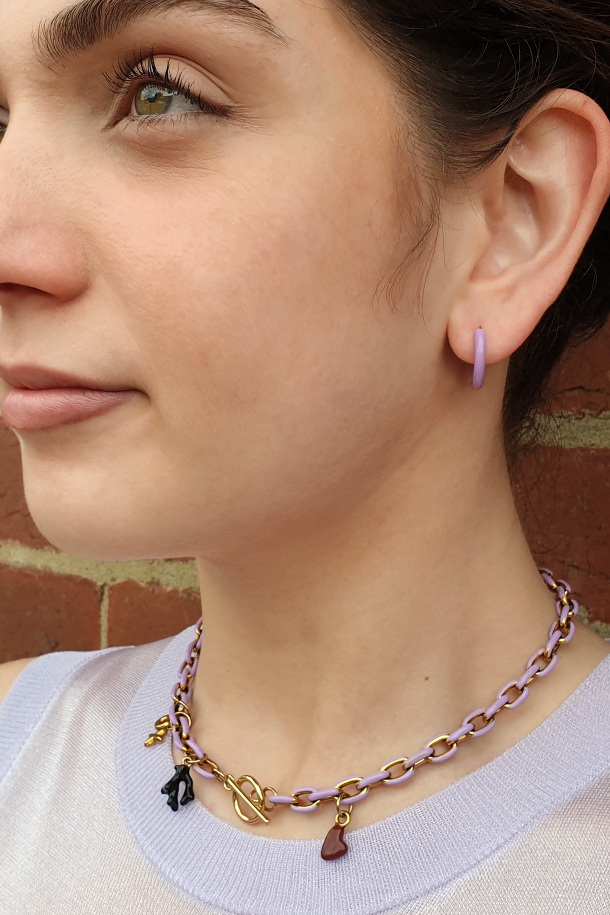 A close-up of a woman wearing a lavender and gold chain necklace with small charms, including a heart and coral, against a brick wall background.