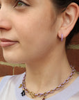 A close-up of a woman wearing a lavender and gold chain necklace with small charms, including a heart and coral, against a brick wall background.