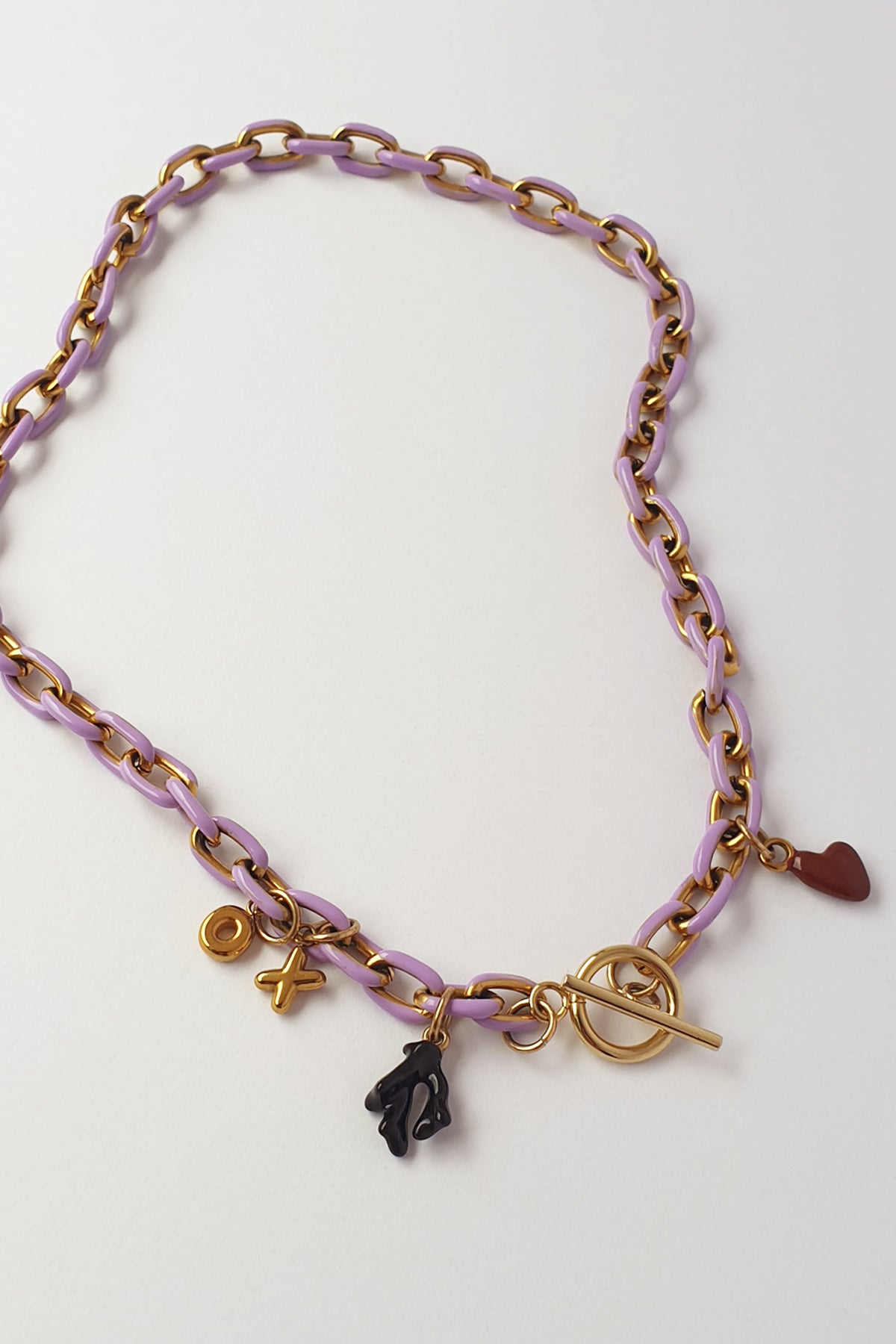 A gold-tone chain necklace with lavender purple links, featuring a toggle clasp, a small black coral shaped charm, a burgandy heart charm, displayed on a light off-white background.