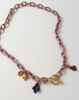 A gold-tone chain necklace with lavender purple links, featuring a toggle clasp, a small black coral shaped charm, a burgandy heart charm, displayed on a light off-white background.