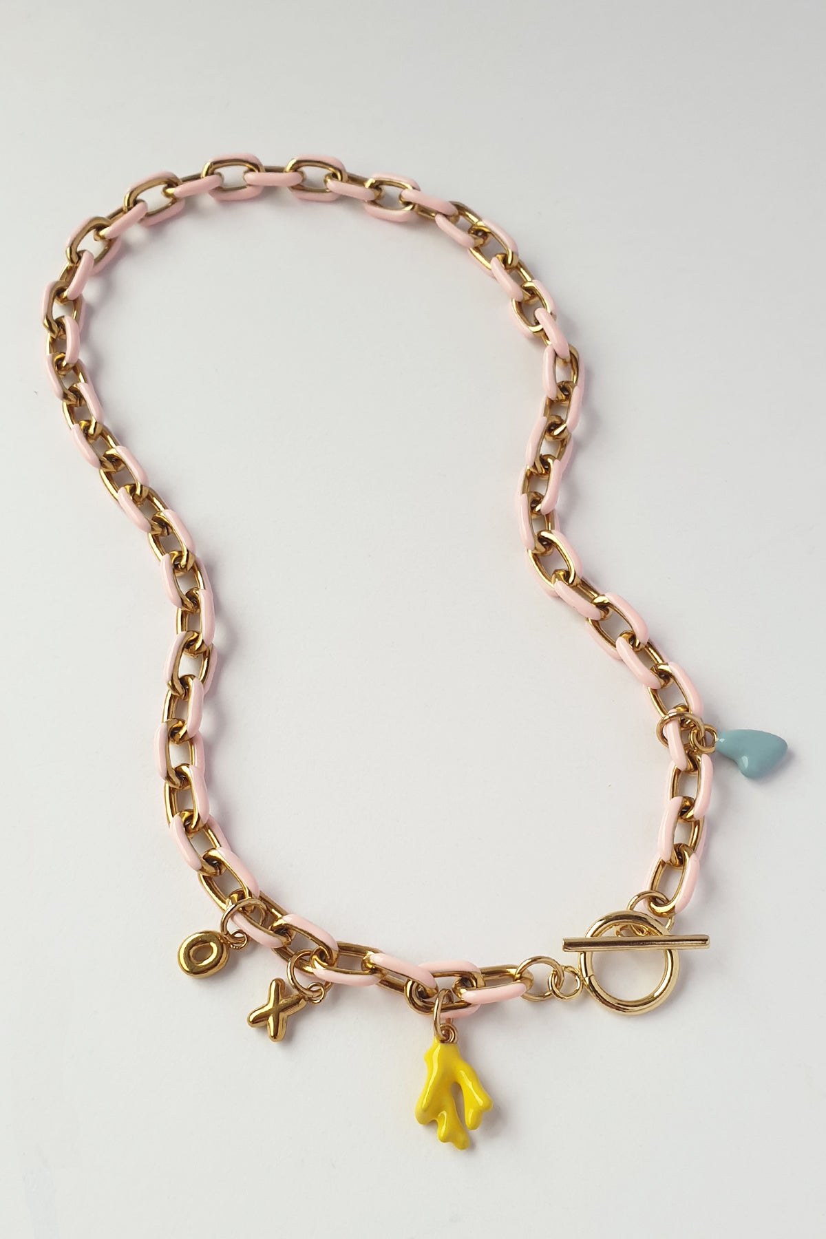 A gold-tone chain necklace with a light pink enamel inlay, featuring small dangling charms including a yellow flower, a light blue heart, and a toggle clasp, displayed on a clean white background.