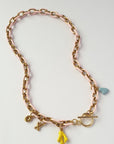 A gold-tone chain necklace with a light pink enamel inlay, featuring small dangling charms including a yellow flower, a light blue heart, and a toggle clasp, displayed on a clean white background.
