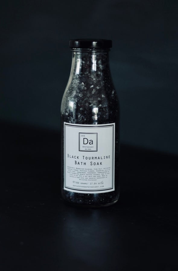 A glass jar filled with bath crytstals and petals sits against a black background. It has a white label with a black border and inside the text Black Tourmaline Bath Soak is written along with a list of ingredients.