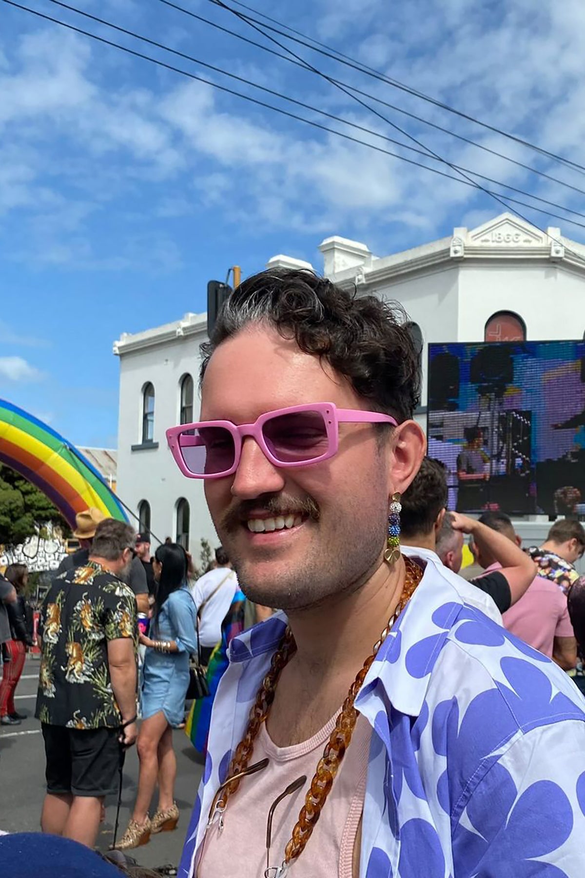 Connor wears the Mirror Mirror earrings at an outdoor event.  He is also wearing pink sunglasses and a white and purple floral shirt over a light pink singlet.  A crowd of people are watching a big screen and an inflatable rainbow is in the background.