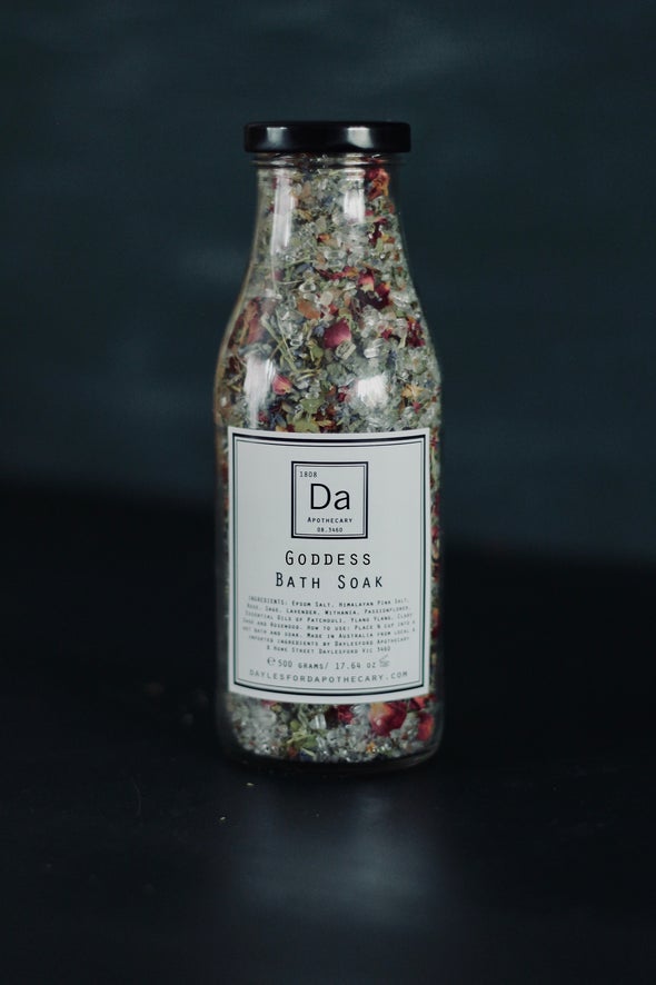 A glass jar filled with bath crytstals and petals sits against a black background. It has a white label with a black border and inside the text Goddess Bath Soak is written along with a list of ingredients. 