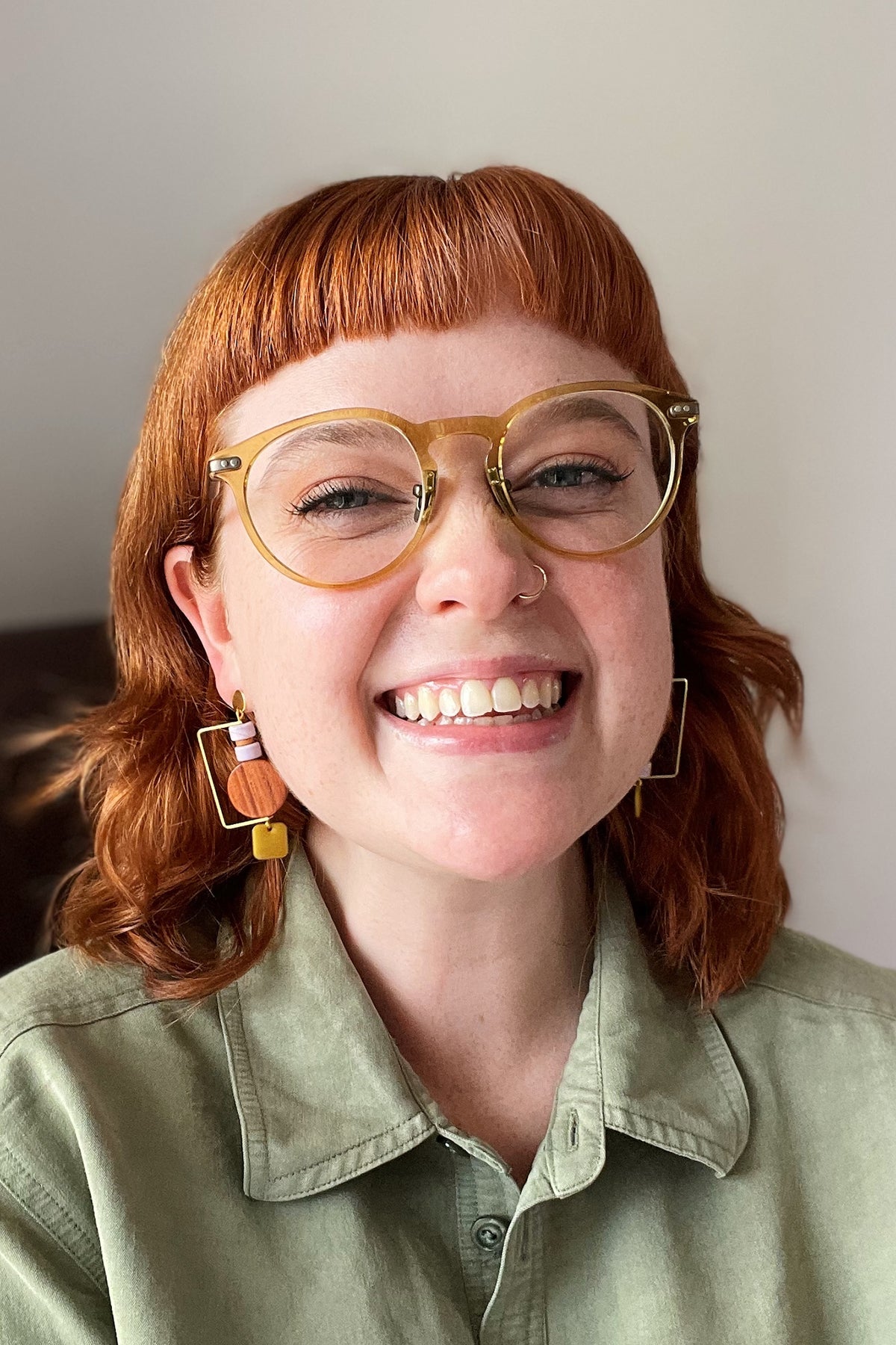 A smiling lady with red hair and yellow glasses stands against a white background. She wears a green linen shirt and a pair of the exhibit earrings in soft mauve and chartreuse.