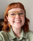 A smiling lady with red hair and yellow glasses stands against a white background. She wears a green linen shirt and a pair of the exhibit earrings in soft mauve and chartreuse.