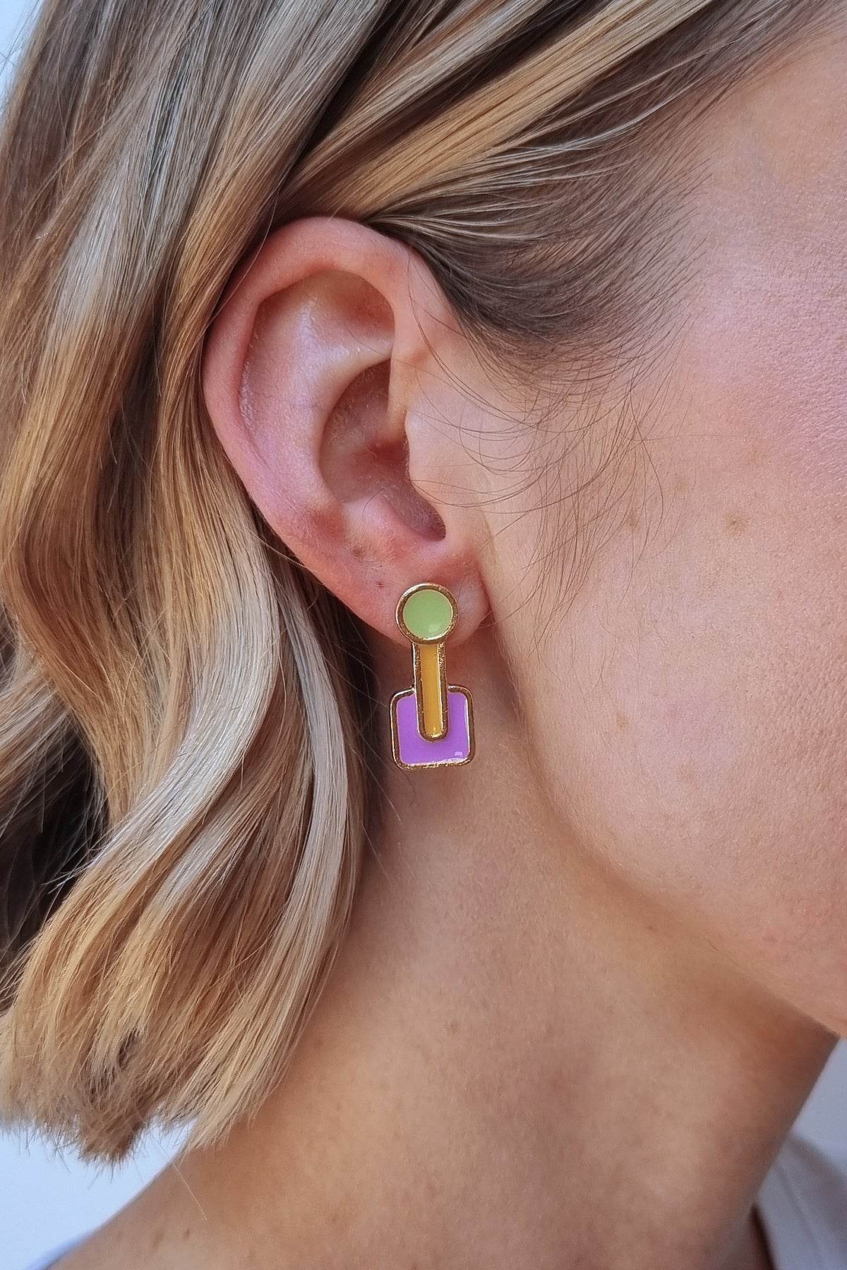 A blonde lady wears the Joystick studs in violet. They feature a green circle up top, a chartreuse drop which ends in the centre of a violet rounded square shape. The shapes are all outlined in gold.