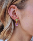 A blonde lady wears the Joystick studs in violet. They feature a green circle up top, a chartreuse drop which ends in the centre of a violet rounded square shape. The shapes are all outlined in gold.