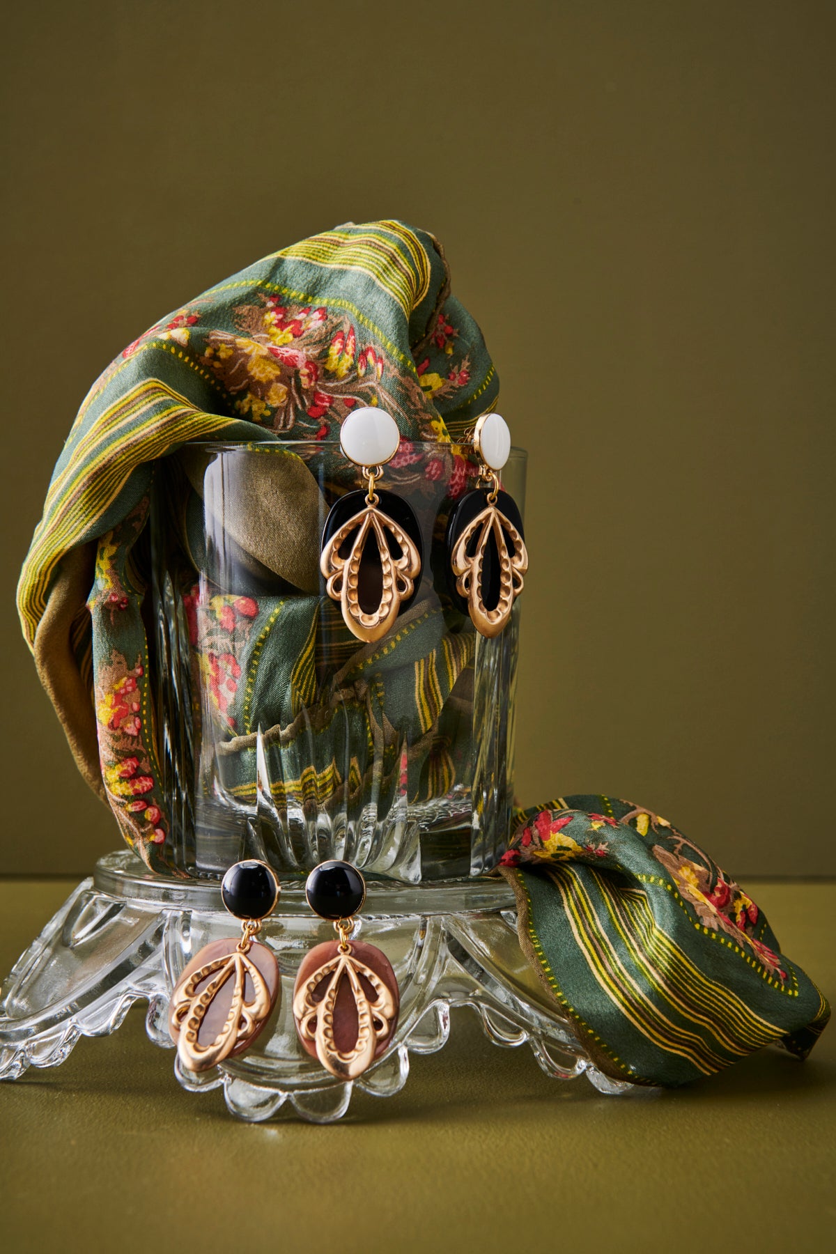 A glass vessel sits styled against an olive green background with a floral scarf pouring out of the vessel. The Dressage earrings are shown hanging from the vessel in both the black and brown colourways