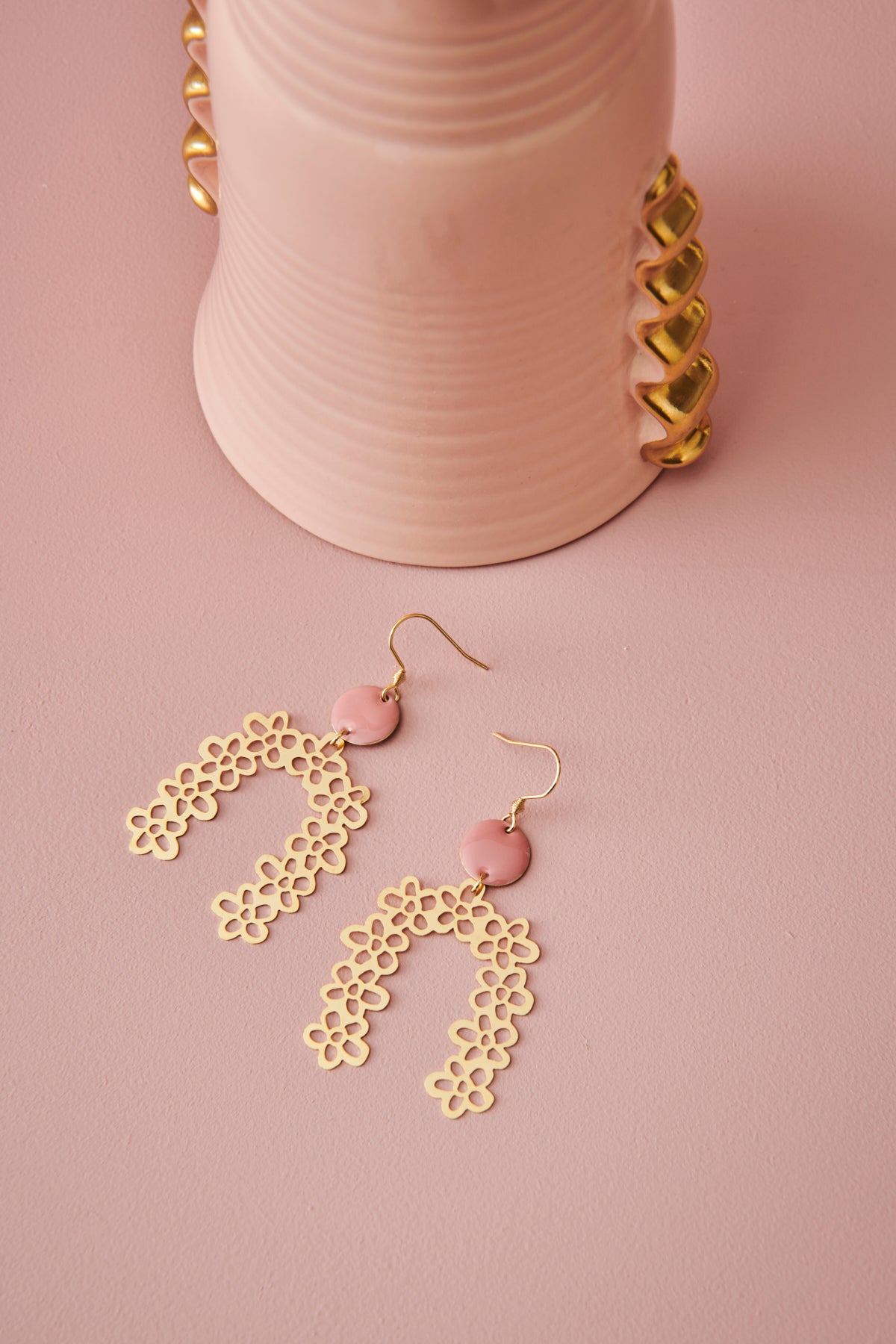 A pair of Drapery earrings in pink sit styled against a pink background. A pink vessel with gold detailing sits nearby.
