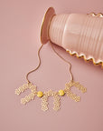 A yellow Drapery necklace sits styled against a pink background with the chain looped over the base of a pink vessel that has gold details.