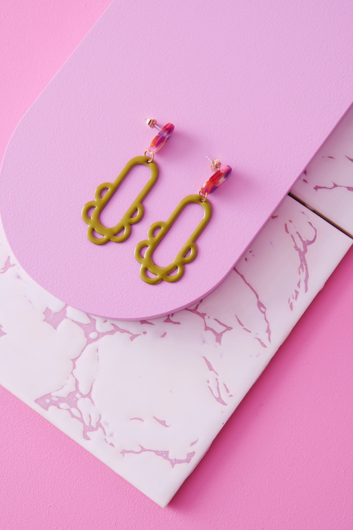 A pair of Fiesta earrings in avocado sit styled against a pink background.