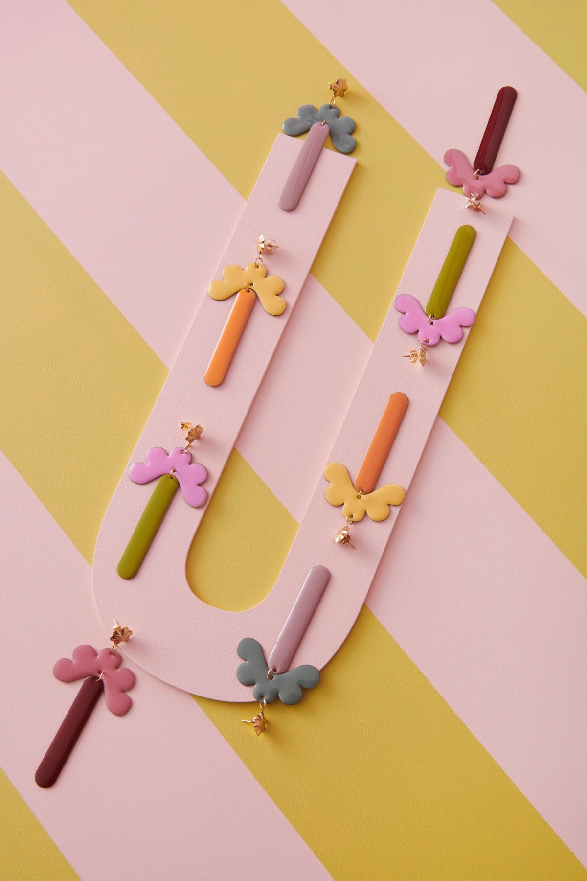 The Meadow earrings in the four colourways; duckegg and mauve, yellow and pumpkin, orchid and avocado, and pink and coffee. are styled against a yellow and pink striped background. The earrings are lined in two rows along a pink U shaped board.