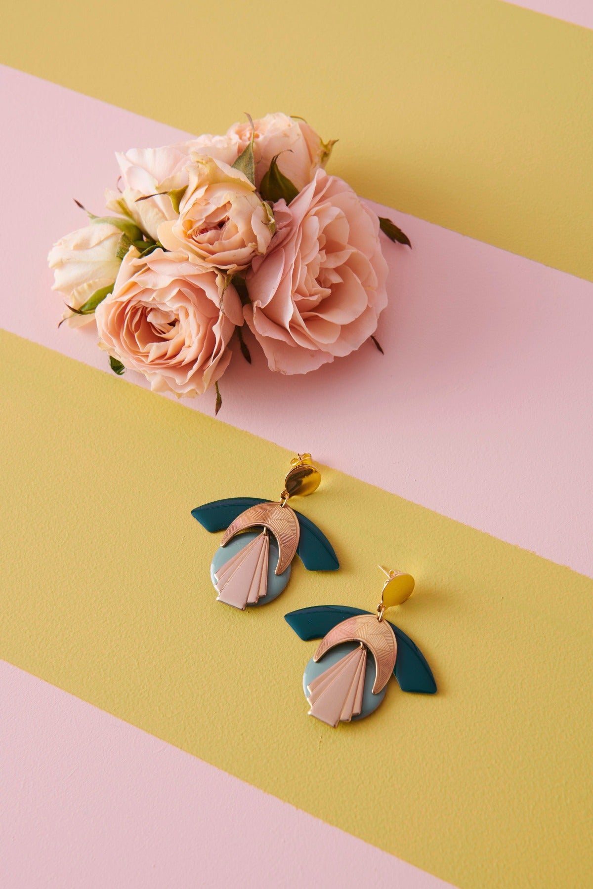 A pair of Diety earrings in the mallard and duckegg colourway lay styled against a yellow and pink striped background. A posy of pink roses lays next to them.