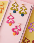 Two pairs of the Fondue earrings in avocado and magenta are styled against a pink board with fondue forks on either side. The pink board sits on a yellow and pink striped background and the chocolate brown, violet and orange Fondue earrings are placed around the edge of the pink board.