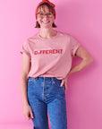 A smiling lady with short dark hair, glasses and a red bow tied around her head wears blue jeans and a rose pink t-shirt that reads just a little bit different. She stands against a bright pink background with her left hand on her hip.