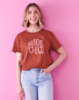 A smiling lady with short wavy blonde hair wears blue jeans and a clay brown t-shirt that reads middle child club. She stands against a bright pink background and has her left hand in her pocket.