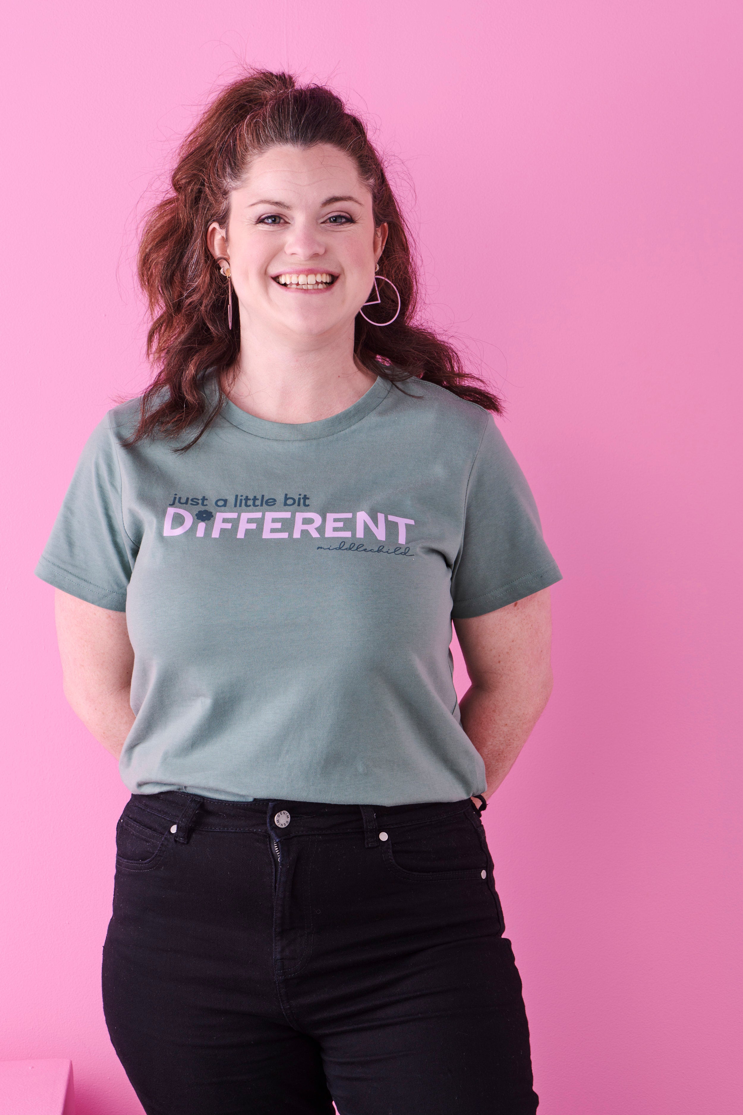 A smiling lady with long wavy dark hair wears black jeans and a sage green t-shirt that reads just a little bit different. She stands against a bright pink background.