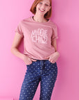 A smiling lady with short red hair wears blue jeans with polkadots and a rose pink t-shirt that reads middle child club. She stands against a bright pink background and has both of her hand in her pockets.