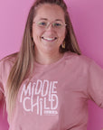 A smiling lady with long blonde hair and glasses wears a rose pink t-shirt that reads middle child club. She stands against a bright pink background and has her left hand on her hip.