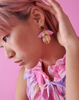 A lady with pink hair sits against a pink background with her hand tucking her hair behind her ear. She models a side view of the Diety earrings in the orchid and peach colour way, and wears a pink floral top.