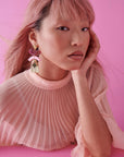 A lady with pink hair sits against a pink background. She models a side view of the Lustre earrings in pink. She wears a pleated pink sheer top and has a hand resting against her face.