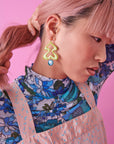 A lady with pink hair holds her hair away from her face to reveal a pair of Fondue earrings in avocado green. She wears a blue retro floral print turtle neck top and a pink pinafore top with blue speckles. She stands against a pink background.