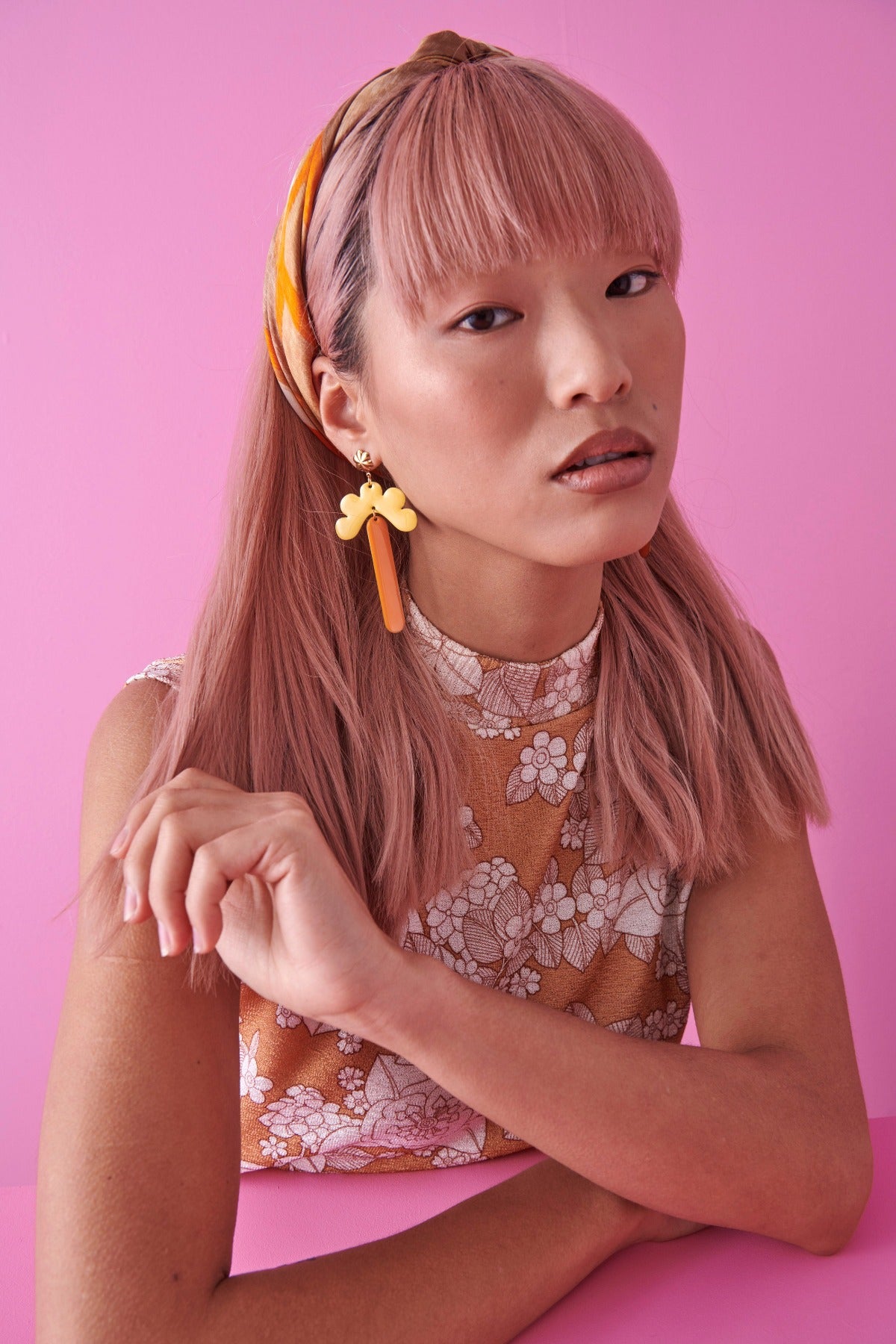 A lady with pink hair sits against a pink background. She models the Meadow earrings in yellow and pumpkin. She wears a mustard and cream retro floral top and has an orange scarf tied through her hair.